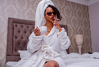 A black woman wearing a white robe sitting in bed holding a cup of coffee in one hand and a cigarette in the other.